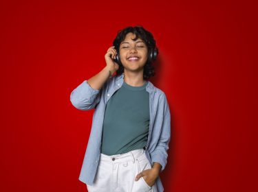 Auf dem Artikel-Bild zu Songs aus der Vodafone Werbung steht eine Frau mit geschlossenen Augen vor rotem Hintergrund, trägt schwarze Kopfhörer und hört Musik.