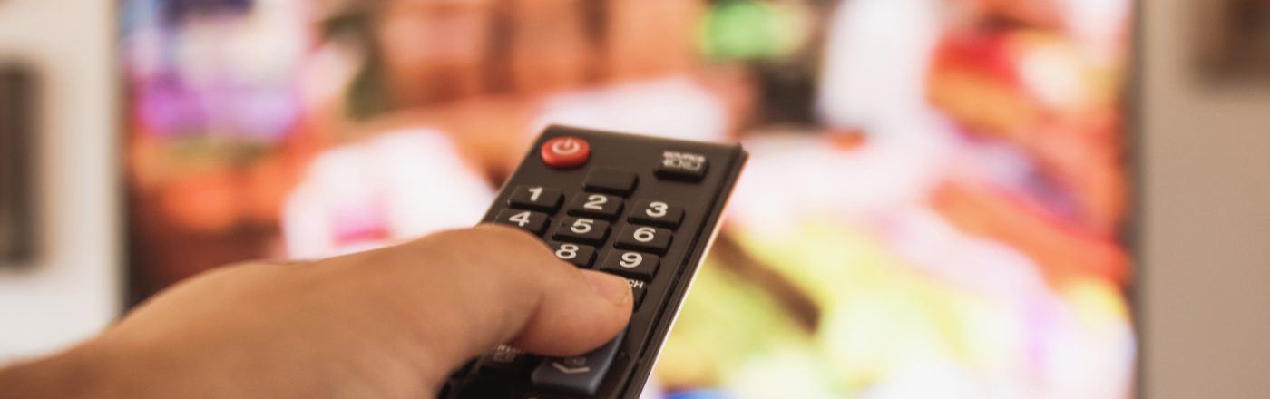 Eine Hand hält eine Fernbedienung, während im Hintergrund ein unscharfer Fernseher mit bunten Inhalten zu sehen ist. Dieses Bild illustriert das Streaming-Erlebnis, passend zum Thema Crunchyroll Kosten 2025.