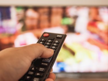 Eine Hand hält eine Fernbedienung, während im Hintergrund ein unscharfer Fernseher mit bunten Inhalten zu sehen ist. Dieses Bild illustriert das Streaming-Erlebnis, passend zum Thema Crunchyroll Kosten 2025.