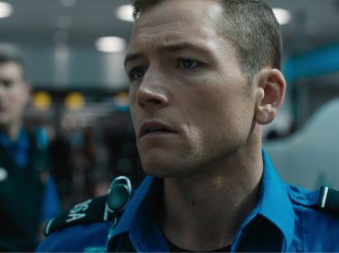 Auf dem Bild aus dem Film Carry-On ist Taron Egerton als Ethan Kopek zu sehen. Er trägt eine blaue Uniform mit TSA-Aufschrift und wirkt ernst und konzentriert. Im unscharfen Hintergrund ist eine weitere Person in ähnlicher Uniform zu erkennen, die die Flughafenatmosphäre unterstreicht. Die Szene vermittelt Spannung und Dramatik.