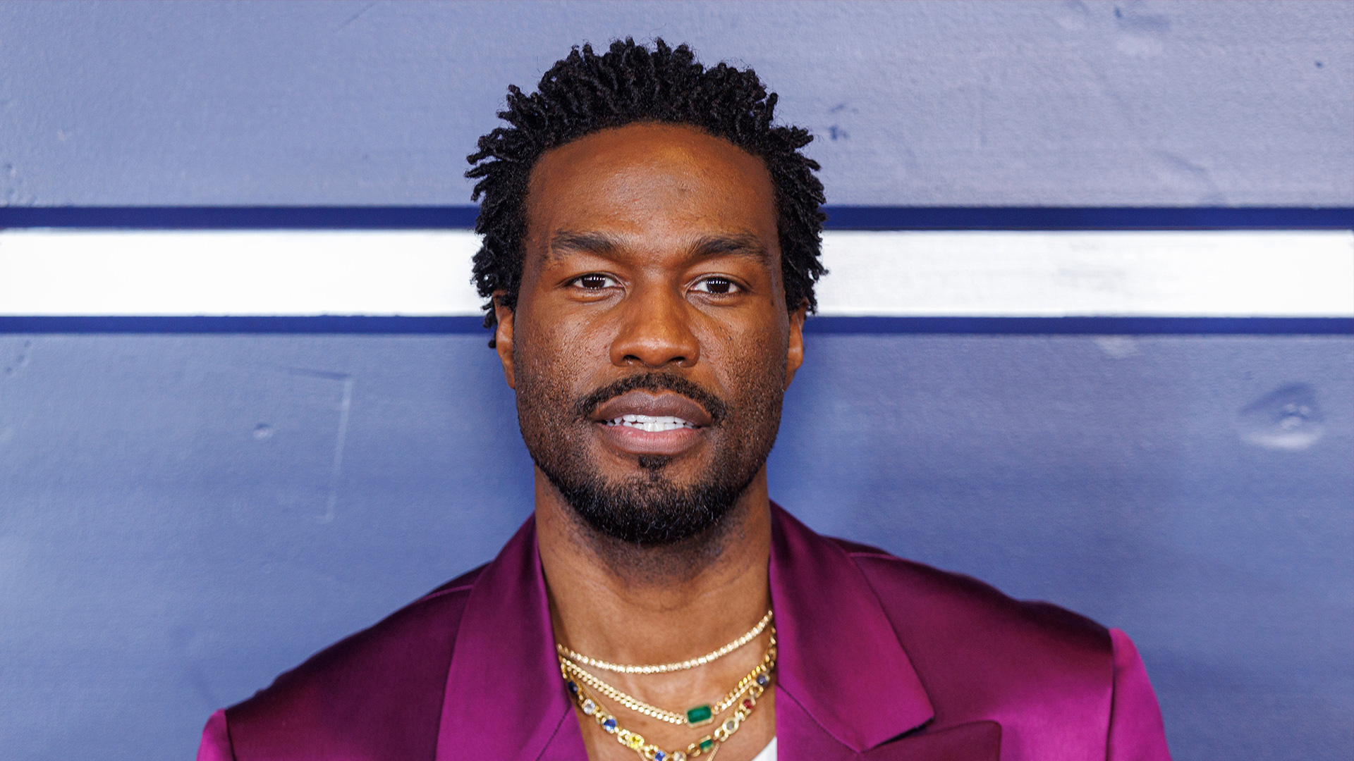 Auf dem Bild zu "Marvels Wonder Man" ist Yahya Abdul-Mateen II zu sehen. Er trägt ein auffälliges, purpurfarbenes Sakko über einem weißen Oberteil mit "Versace"-Schriftzug. Mehrere goldene Halsketten mit Edelsteinen vervollständigen seinen Look. Der Hintergrund ist schlicht blau mit einer weißen Linie, was den Fokus auf sein stilvolles Outfit und seine selbstbewusste Ausstrahlung lenkt.