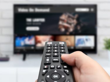 Auf dem Bild in dem Artikel zu den RTL+ Abos 2025 ist eine Hand zu sehen, die eine schwarze Fernbedienung hält und auf einen Flachbildfernseher zeigt. Auf dem Bildschirm wird eine Streaming-Oberfläche mit verschiedenen Inhalten angezeigt. Im Hintergrund befinden sich eine weiße Ziegelwand, ein kleiner Pflanzentopf und eine aufgeräumte, moderne Wohnzimmerumgebung.