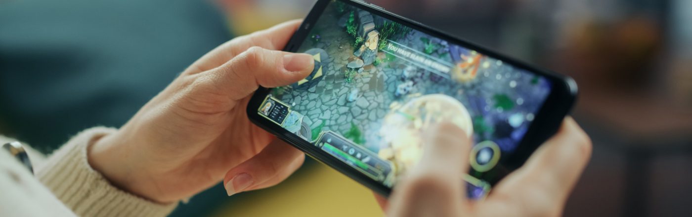 Auf dem Bild ist eine Person zu sehen, die ein Smartphone in beiden Händen hält und ein Strategiespiel spielt. Der Bildschirm zeigt eine farbenfrohe Spielszene mit Figuren und Steinelementen. Die entspannte Umgebung im Hintergrund deutet auf Freizeit hin. Das Bild thematisiert Handyspiele für Erwachsene.