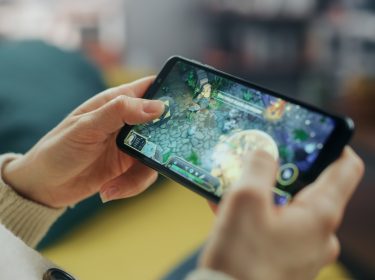 Auf dem Bild ist eine Person zu sehen, die ein Smartphone in beiden Händen hält und ein Strategiespiel spielt. Der Bildschirm zeigt eine farbenfrohe Spielszene mit Figuren und Steinelementen. Die entspannte Umgebung im Hintergrund deutet auf Freizeit hin. Das Bild thematisiert Handyspiele für Erwachsene.