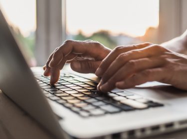 Auf dem Bild in dem Artikel zu den Kosten von ChatGPT im Jahr 2025 sind Hände zu sehen, die auf der Tastatur eines Laptops tippen. Die Szene ist in warmem Tageslicht gehalten, das durch ein Fenster im Hintergrund einfällt, was eine produktive und konzentrierte Atmosphäre vermittelt.