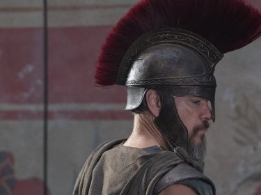 Das Bild aus The Odyssey zeigt Matt Damon in der Rolle des Odysseus. Er trägt eine antike Rüstung mit einem markanten bronzenen Helm, der mit einem roten Kamm verziert ist. Sein Gesichtsausdruck ist ernst und fokussiert, während er zur Seite blickt. Der Hintergrund zeigt eine Wand mit verblassten Fresken, die an antike griechische Kunst erinnern. Die Szene vermittelt eine epische und historische Atmosphäre, passend zur legendären Geschichte von Odysseus.