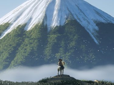 Im Bild aus dem Videospiel Ghost of Yotei steht ein:e Reiter:in mit Hut auf einem Felsen, während sein:ihr Pferd ruhig verweilt. Im Hintergrund erhebt sich ein majestätischer Berg, dessen grüne Wälder von einer Schneeschicht bedeckt sind. Um den Berg fliegen weiße Vögel, die eine friedliche Stimmung unterstreichen. Nebel bedeckt das Tal.