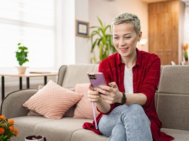 Eine Frau sitzt auf einem Sofa im Wohnzimmer und bedient ein Smartphone.