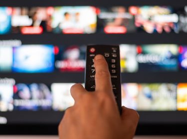 Auf dem Bild in dem Artikel zu den Kosten von Apple TV+ im Jahr 2025 ist eine Hand zu sehen, die eine Fernbedienung hält und auf einen Fernseher zielt. Auf dem Bildschirm sind verschwommene Miniaturansichten von Filmen und Serien in einer Streaming-Oberfläche sichtbar. Die Szene vermittelt eine moderne, digitale Atmosphäre.