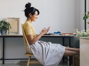 Seitenansicht einer jungen Frau, die am Schreibtisch sitzt und auf ihr Smartphone schaut.
