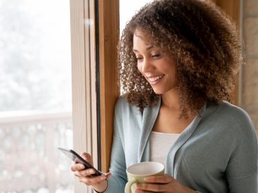 Frau schaut lächelnd auf ihr Android-Smartphone, während sie eine Tasse Tee in der Hand hält.