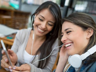 Zwei Frauen teilen sich Kopfhörer und hören gemeinsam Musik mit dem Smartphone.
