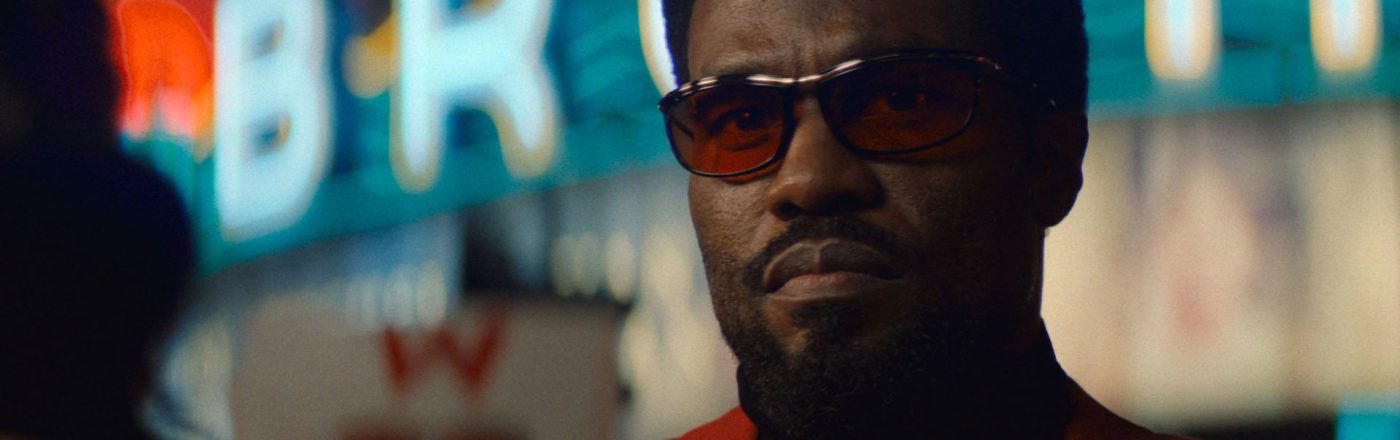 In dieser Szene aus der Wonder Man-Serie ist Yahya Abdul-Mateen II als Simon Williams zu sehen. Er trägt ein auffälliges rotes Sakko, dunkle Sonnenbrille und blickt ernst in die Ferne.