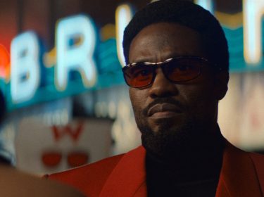 In dieser Szene aus der Wonder Man-Serie ist Yahya Abdul-Mateen II als Simon Williams zu sehen. Er trägt ein auffälliges rotes Sakko, dunkle Sonnenbrille und blickt ernst in die Ferne.