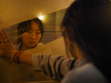 Im Bild zum Until Dawn-Film ist Megan, gespielt von Ji-young Joo, zu sehen. Sie blickt ernst in einen schmutzigen Spiegel, während ihre Hand gegen die spiegelnde Oberfläche drückt. Die Szene ist in warmes, gedämpftes Licht getaucht, was eine nachdenkliche und emotionale Stimmung erzeugt.