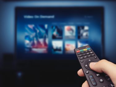 Auf dem Bild in dem Artikel zu den Prime Video-Kosten hält eine Person eine schwarze Fernbedienung in der Hand und navigiert durch das Menü eines Streaming-Dienstes auf einem großen Bildschirm. Die Oberfläche zeigt verschiedene Filme und Serien zur Auswahl. Die Szene ist in dunklen Blau- und Schwarztönen gehalten, was eine gemütliche Heimkino-Atmosphäre erzeugt.