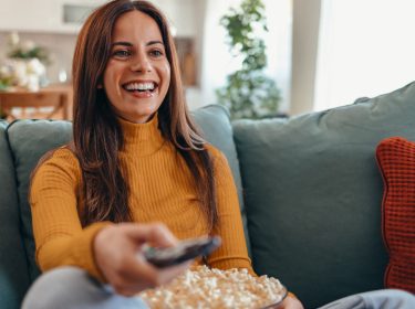 Eine junge Frau sitzt auf dem Sofa und hält eine Fernbedienung in der Hand.
