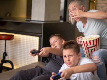 Zwei Jungen sitzen auf einem Sofa und spielen Mario Kart 9 auf der Nintendo Switch 2. Beide halten Controller und wirken konzentriert, während eine Frau hinter ihnen sitzt, Popcorn aus einer großen, rot-weiß gestreiften Schüssel hält und lachend mit der Hand ihr Gesicht teilweise bedeckt. Die Stimmung ist lebendig und entspannt, passend zu einer fröhlichen Spielrunde.