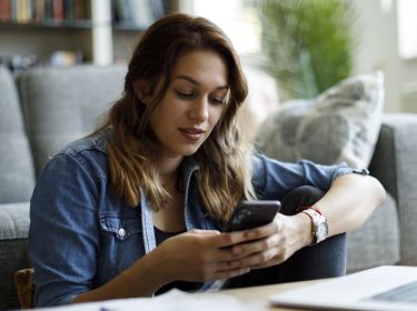 Junge Frau sitzt auf dem Boden vor ihrem Sofa und schaut lächelnd auf ihr Android-Handy.