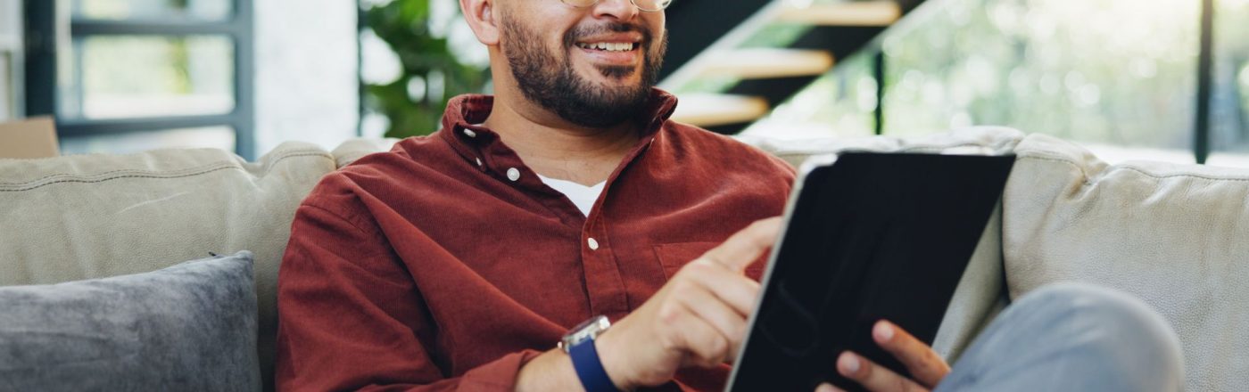 Junger Mann sitzt auf der Couch und arbeitet an seinem Tablet.