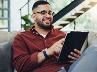 Junger Mann sitzt auf der Couch und arbeitet an seinem Tablet.