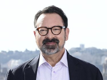 Auf dem Bild in dem Artikel zu Dawn of the Jedi ist James Mangold zu sehen. Er trägt eine dunkle Brille, hat einen gepflegten Bart und trägt ein helles Hemd mit einem dunklen Sakko. Im Hintergrund erstreckt sich eine Stadtlandschaft mit historischen Gebäuden und Kuppeln. Mangold lächelt leicht und blickt direkt in die Kamera, was eine freundliche und selbstbewusste Ausstrahlung vermittelt.