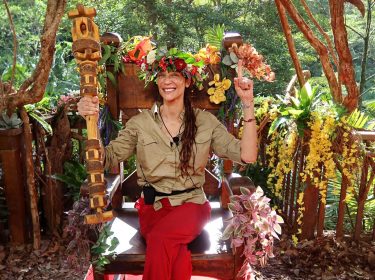 Auf dem Bild zum Dschungelcamp 2025 ist Lilly Becker als frisch gekrönte Dschungelkönigin zu sehen. Sie sitzt auf einem hölzernen Thron, trägt eine bunte Blumenkrone und hält das Siegerzepter in der Hand. Mit einem strahlenden Lächeln und erhobenem Finger feiert sie ihren Sieg. Der Thron ist mit bunten Blumen und Pflanzen geschmückt, im Hintergrund ist dichter, grüner Dschungel zu sehen. Die Stimmung ist feierlich und triumphierend.