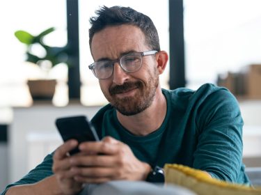 Ein Mann sitzt auf einem Sofa und schaut auf ein Smartphone, das er in seinen Händen hält