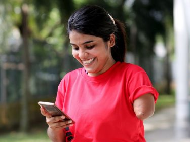Junge Frau schaut lächelnd auf ein Smartphone, das sie in der rechten Hand hält.