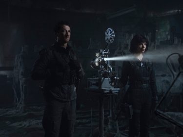 Auf dem Bild zu The Gorge Ende erklärt sind Anya Taylor-Joy als Drasa und Miles Teller als Levi in einer düsteren, verfallenen Umgebung zu sehen. Die beiden tragen taktische schwarze Kleidung und stehen vor einem alten Filmprojektor, der einen Lichtstrahl durch den staubigen Raum wirft. Levi blickt ernst in die Ferne, während Drasa konzentriert auf das projizierte Bild schaut. Die Szenerie wirkt geheimnisvoll und angespannt, verstärkt durch die spärliche Beleuchtung und die unheimliche Atmosphäre des verlassenen Ortes.