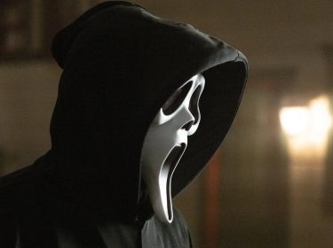 Auf dem Bild zu Scream 7 ist der ikonische Killer Ghost Face zu sehen. Er trägt eine schwarze Kapuze und die bekannte weiße Maske mit weit aufgerissenem Mund und leeren Augen. Die Szene wirkt düster und unheimlich, mit verschwommenen Lichtern im Hintergrund, die eine bedrohliche Atmosphäre erzeugen