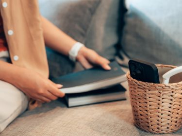 Eine Frau legt ihr Smartphone in einen kleinen geflochtenen Korb, der neben weißen Kopfhörern auf einem Sofa steht. Stattdessen hält sie ein Buch in der Hand, bereit zu lesen. Die Szene symbolisiert das Konzept 