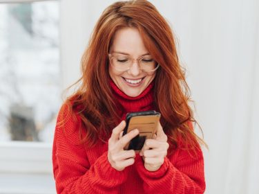 Rothaarige junge Frau in rotem Pullover schaut auf Android-Handy, das sie in den Händen hält.
