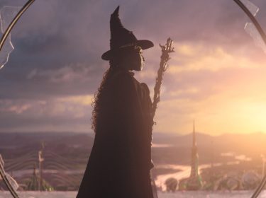 Auf dem Bild in dem Artikel zu den Oscar Filmen 2025, die bei GigaTV verfügbar sind, ist eine Szene aus Wicked: Teil 1 zu sehen. Eine Frau mit grüner Haut steht in einem zerbrochenen Fensterrahmen und blickt in die Ferne. Sie trägt einen spitzen Hexenhut und einen langen schwarzen Umhang. Die untergehende Sonne taucht die Landschaft in goldenes Licht, während eine Stadt mit hohen Türmen im Hintergrund liegt. Die Szene wirkt episch und mystisch.