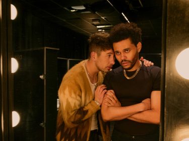 Auf dem Bild aus Hurry Up Tomorrow Film sind Barry Keoghan und Abel Tesfaye (The Weeknd) in einer intensiven Szene zu sehen. Barry Keoghan, in einer gemusterten Jacke, lehnt sich nah an Tesfaye heran und flüstert ihm etwas zu, während er eine ernste Miene zeigt. Abel Tesfaye steht mit verschränkten Armen vor einem Spiegel mit hellen Glühbirnen und wirkt angespannt. Die dunkle Garderobenraum-Atmosphäre verstärkt die dramatische Stimmung.