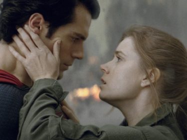 Auf dem Bild zu Superman-Filme Reihenfolge sind Henry Cavill als Superman und Amy Adams als Lois Lane in einer emotionalen Szene zu sehen. Superman trägt sein ikonisches blau-rotes Kostüm, während Lois in einer olivgrünen Jacke gekleidet ist. Sie hält sein Gesicht mit beiden Händen, und beide blicken sich intensiv in die Augen. Im Hintergrund sind Feuer und Rauch zu sehen, was auf eine dramatische Situation hindeutet.