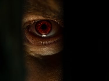 Ein unheimliches, rot leuchtendes Auge blickt durch einen schmalen Spalt. Die düstere Beleuchtung und der intensive Blick erzeugen eine bedrohliche Atmosphäre, die an Szenen aus Monsterfilmen erinnert. Der Fokus liegt auf dem Auge, das fast übernatürlich wirkt.