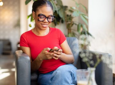 Lächelnde Frau mit roten T-Shirt schaut lächelnd auf ihr Smartphone, das sie in den Händen hält.