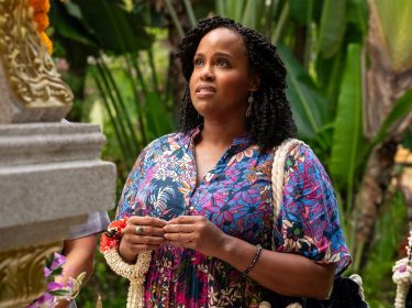 Auf dem Bild aus The White Lotus Staffel 3 ist Natasha Rothwell in Episode 1 als Belinda Lindsey zu sehen. Sie trägt ein farbenfrohes, blumiges Kleid und blickt nachdenklich auf eine verzierte Steinstruktur. In ihren Händen hält sie Blumenschmuck. Der Hintergrund ist von tropischen Pflanzen und traditionellen Dekorationen geprägt, was die exotische Atmosphäre unterstreicht. Der The White Lotus Staffel 3 Cast umfasst unter anderem ihre Rolle.