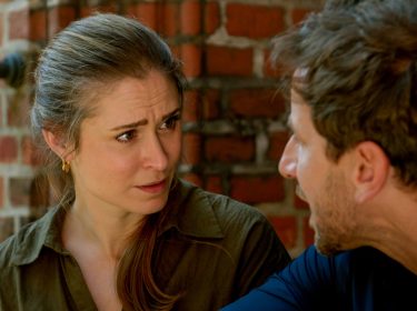 Auf dem Bild aus Rote Rosen sind die Darsteller:innen Lea Marlen Woitack als Svenja und Vivian Frey als Arthur zu sehen. Die Szene zeigt ein intensives Gespräch zwischen den beiden. Svenja blickt Arthur mit ernster Miene und leicht besorgtem Ausdruck an, während er spricht. Der Hintergrund mit rotem Ziegelmauerwerk verleiht der Szene eine warme, aber zugleich angespannte Atmosphäre.