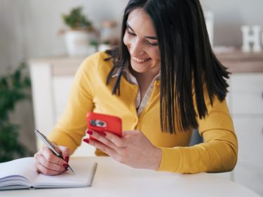 Eine Frau hält ein Smartphone in der Hand und macht nebenbei Notizen in einem Kalender.