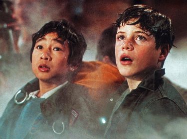 Auf dem Bild in dem Artikel zur Die Goonies-Fortsetzung sind Ke Huy Quan als Data und Sean Astin als Mikey in einer spannenden Szene zu sehen. Beide blicken überrascht und ehrfürchtig in die Ferne, während dichter Nebel die Umgebung umhüllt. Data trägt eine Jacke mit Riemen, während Mikey in einem dunklen Hemd zu sehen ist. Die Szene strahlt Abenteuer und Entdeckung aus.