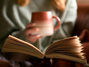 Nachaufnahme eines aufgeschlagenen Buchs, dahinert eine Frau mit einer Tasse.