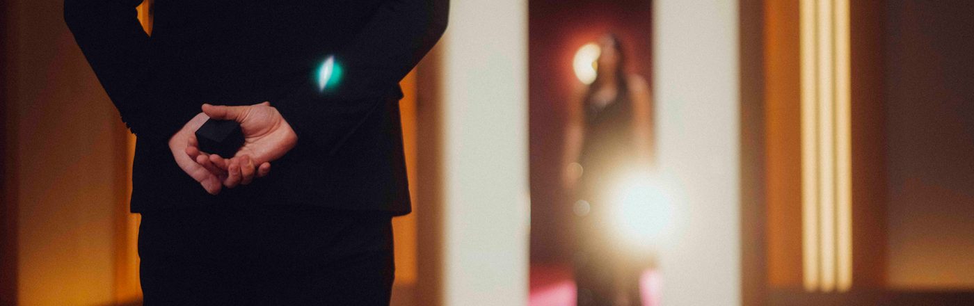 Auf dem Bild in dem Artikel zu Love Is Blind: Germany Staffel 2 steht ein Mann im schwarzen Anzug mit dem Rücken zur Kamera auf einem roten Teppich und hält eine kleine schwarze Schachtel hinter dem Rücken. Im Hintergrund erscheint eine Frau im Abendkleid.