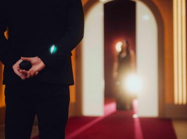 Auf dem Bild in dem Artikel zu Love Is Blind: Germany Staffel 2 steht ein Mann im schwarzen Anzug mit dem Rücken zur Kamera auf einem roten Teppich und hält eine kleine schwarze Schachtel hinter dem Rücken. Im Hintergrund erscheint eine Frau im Abendkleid.