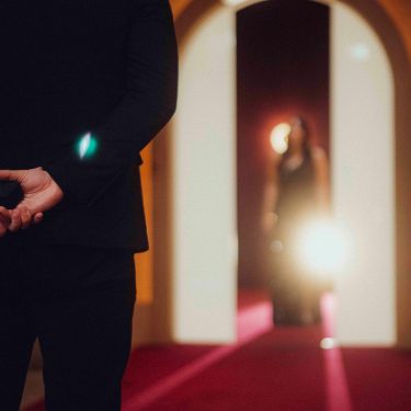 Auf dem Bild in dem Artikel zu Love Is Blind: Germany Staffel 2 steht ein Mann im schwarzen Anzug mit dem Rücken zur Kamera auf einem roten Teppich und hält eine kleine schwarze Schachtel hinter dem Rücken. Im Hintergrund erscheint eine Frau im Abendkleid.