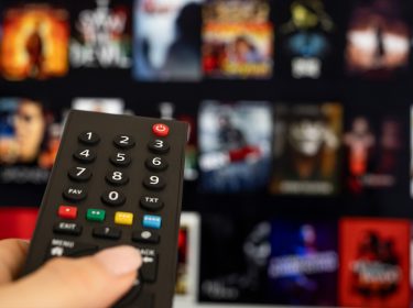 Auf dem Bild ist eine Hand zu sehen, die eine schwarze Fernbedienung hält und auf einen Fernseher zeigt. Der Bildschirm im Hintergrund zeigt die Benutzeroberfläche eines Streaming-Dienstes mit verschiedenen Film- und Serien-Covern, die leicht unscharf sind. Das Bild vermittelt eine typische Streaming-Situation und passt zum Thema Heimkino. Paramount+ könnte als einer der verfügbaren Dienste dargestellt sein.