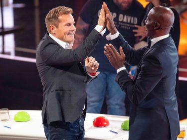 Auf dem Bild in dem Artikel zu den Supertalent-Gewinner:innen sind zwei Juroren zu sehen. Der linke Juror, ein Mann mit kurzen blonden Haaren, trägt ein schwarzes Sakko und ein weißes Hemd, während er lächelnd die Hand hebt. Der rechte Juror, ein Mann mit Glatze im dunklen Anzug, erwidert das High-Five mit begeisterter Mimik. Im Hintergrund ist die Bühne mit leuchtenden Farben und Zuschauer:innen zu erkennen.