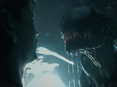 Auf dem Bild zu Alien: Romulus steht eine Person in angespannter Konfrontation mit einem Xenomorph. Das ikonische Alien, mit glänzendem, biomechanischem Kopf und tropfendem Speichel, nähert sich bedrohlich. Die dunkle, neblige Atmosphäre und das kalte Licht im Hintergrund verstärken die Spannung. Die angedeutete Silhouette der Person unterstreicht die Gefahr und das Gefühl von Ausweglosigkeit in dieser intensiven Szene.