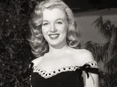 Auf dem Bild, das symbolisch für Dokus über Frauen zum Weltfrauentag verwendet wird, ist Marilyn Monroe in einer eleganten Pose zu sehen. Sie trägt ein schwarzes, schulterfreies Kleid mit Spitzenverzierung und Schleifen. Ihr blondes Haar fällt in sanften Wellen, und sie strahlt mit einem freundlichen Lächeln. Der Hintergrund ist unscharf und zeigt Pflanzen, was das Bild natürlich wirken lässt.